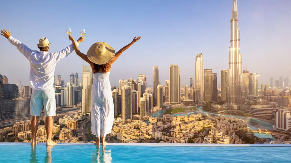 A happy tourist couple on vacation time stands by the pool edge and enjoys the panoramic sunset view of the Dubai city skyline, UAE