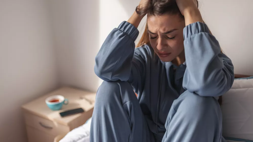 Anxious young woman wearing pajamas, sitting on bed in the morning, holding head in hands, crying