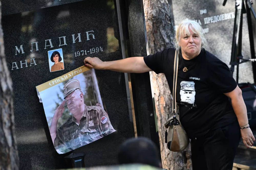 A woman holds a poster of the former Bosnian Serb army commander Ratko Mladic, who died last week while serving a life sentence for genocide and other atrocities during the war in Bosnia, against the grave stone of his daughter Ana Mladic, at the Topcider Cemetery, in Belgrade, Serbia, Monday, Sept. 7, 2026. Text reads "Serbian Hero". (AP Photo)