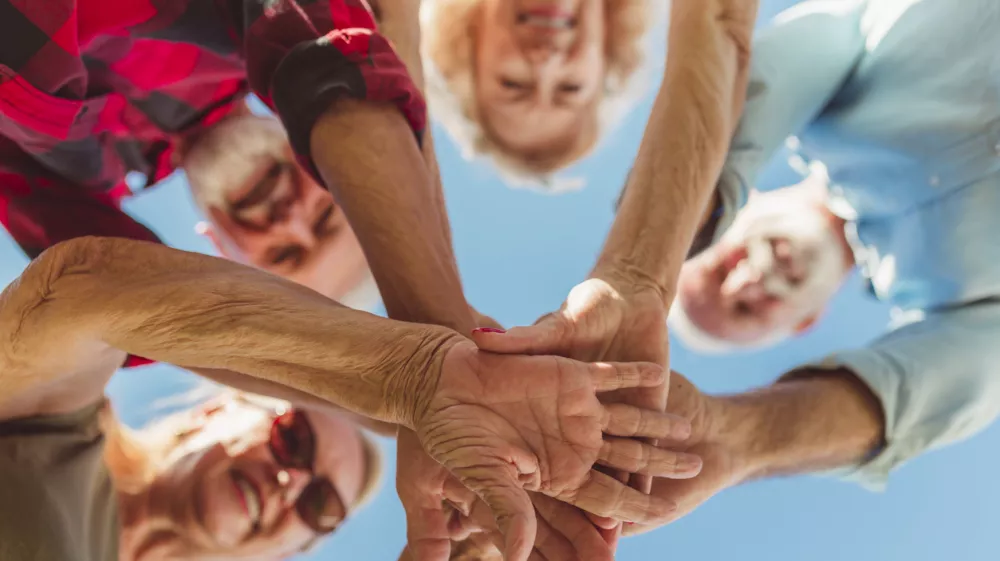 Low angle view of group of cheerful active senior people enjoying sunny summer day outdoors, gathered in circle, holding hands all together in the middle / Foto: Vladans Getty Images/istockphoto