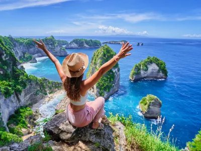 Woman traveler with open arms sitting on rock cliff and enjoying of scenic landscape of Diamond beach during vacation to Nusa Penida Island in Indonesia, travel to touristic beautiful destinations