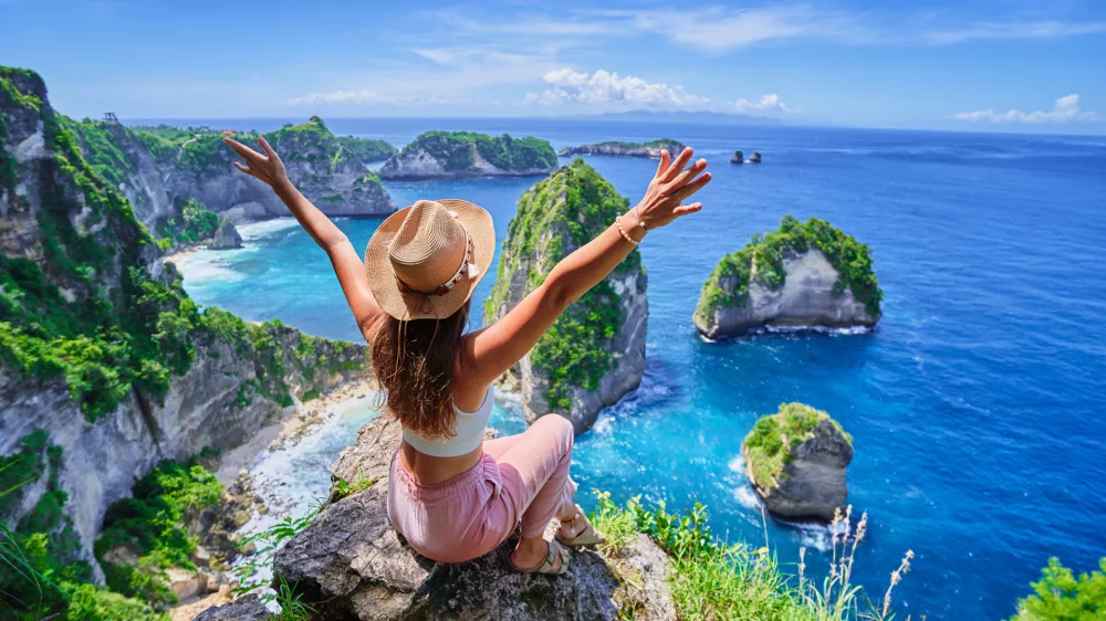 Woman traveler with open arms sitting on rock cliff and enjoying of scenic landscape of Diamond beach during vacation to Nusa Penida Island in Indonesia, travel to touristic beautiful destinations