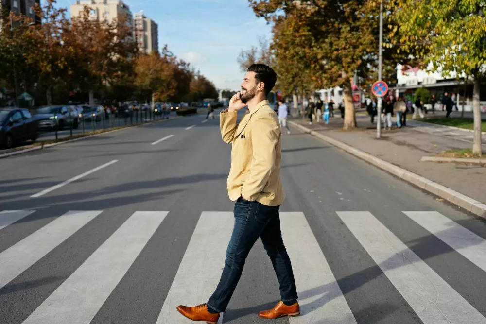 A professional business person in smart casual clothing walks across a zebra crossing while engaging in a mobile phone conversation during a city walk. / Foto: Duxx