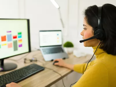 Latin freelancer wearing wireless headset typing on laptop, managing remote project and providing customer support from home workspace