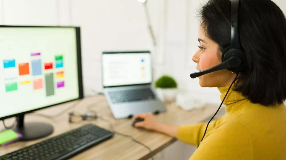 Latin freelancer wearing wireless headset typing on laptop, managing remote project and providing customer support from home workspace