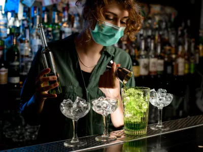 Young bartender girl in a medical mask pour green liquid from beaker into glass with ice. Medical mask for prophylaxis and protection from coronavirus COVID-19.