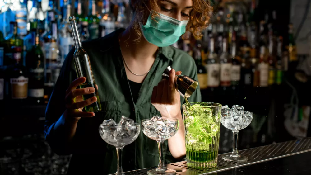 Young bartender girl in a medical mask pour green liquid from beaker into glass with ice. Medical mask for prophylaxis and protection from coronavirus COVID-19.