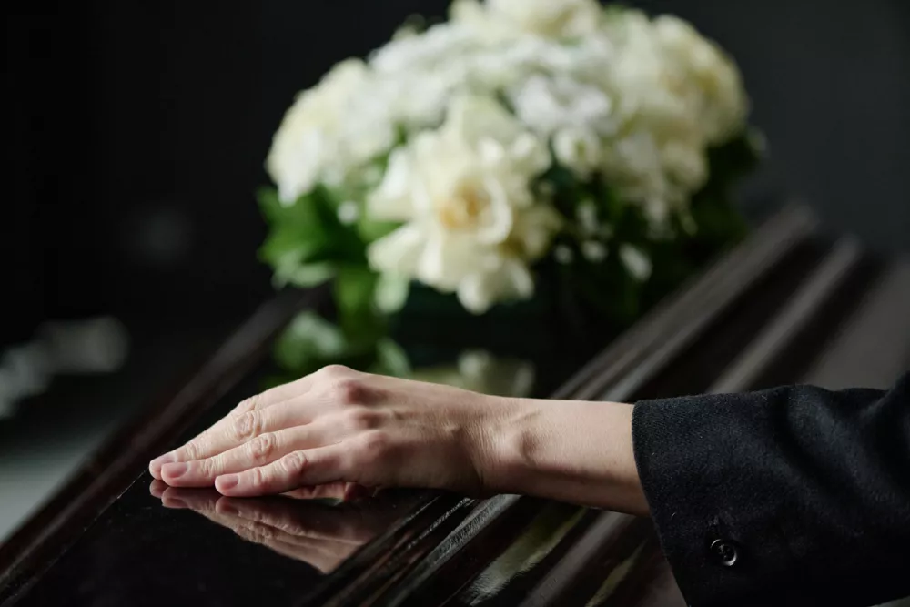Caucasian middle aged woman resting hand on closed coffin during funeral ceremony, blurred bouquet of flowers in background, expressing grief and loss / Foto: Annastills Getty Images