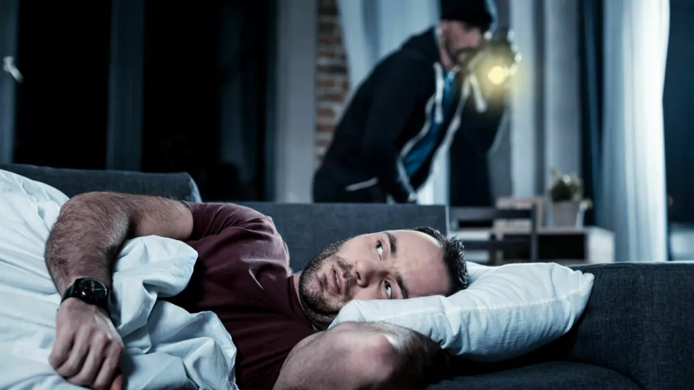 Breaking into house. Alarmed disturbed bearded man waking up and hearing someone being in his house and a criminal with a torch standing behind him / Foto: Zinkevych