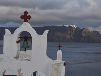 A bell towel of an Orthodox church in the town of Oia on the earthquake-struck island of Santorini, Greece, as the main town of Fira is litted by the sun in the background on Tuesday, Feb. 4, 2025. (AP Photo/Petros Giannakouris)