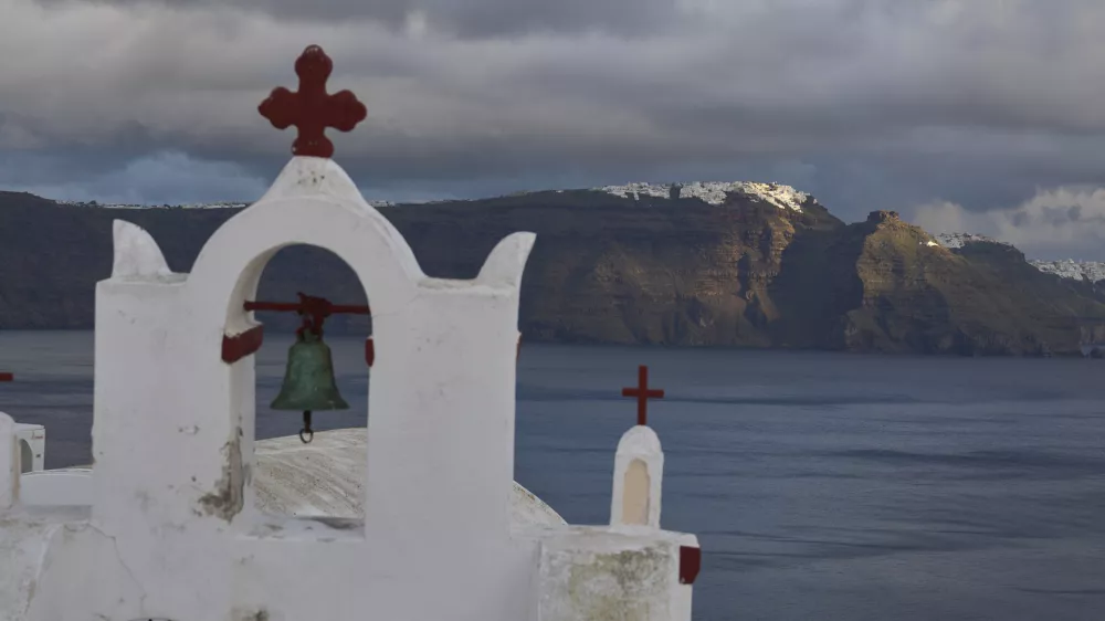 A bell towel of an Orthodox church in the town of Oia on the earthquake-struck island of Santorini, Greece, as the main town of Fira is litted by the sun in the background on Tuesday, Feb. 4, 2025. (AP Photo/Petros Giannakouris)