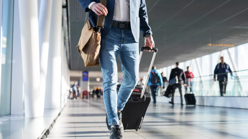 Man with shoulder bag and hand luggage walking in airport terminal, photo with men's legs walking in airport terminal