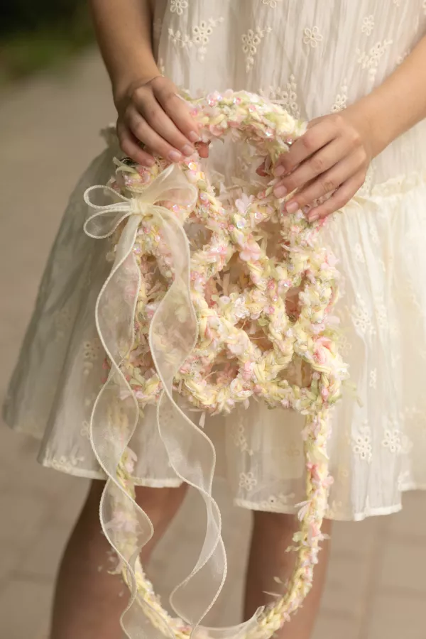 Close-up portrait of young girl holding pastel floral braided wreath with sequins and long sheer ribbon, wearing cream embroidered dress with delicate texture, gentle hands at waist level, soft