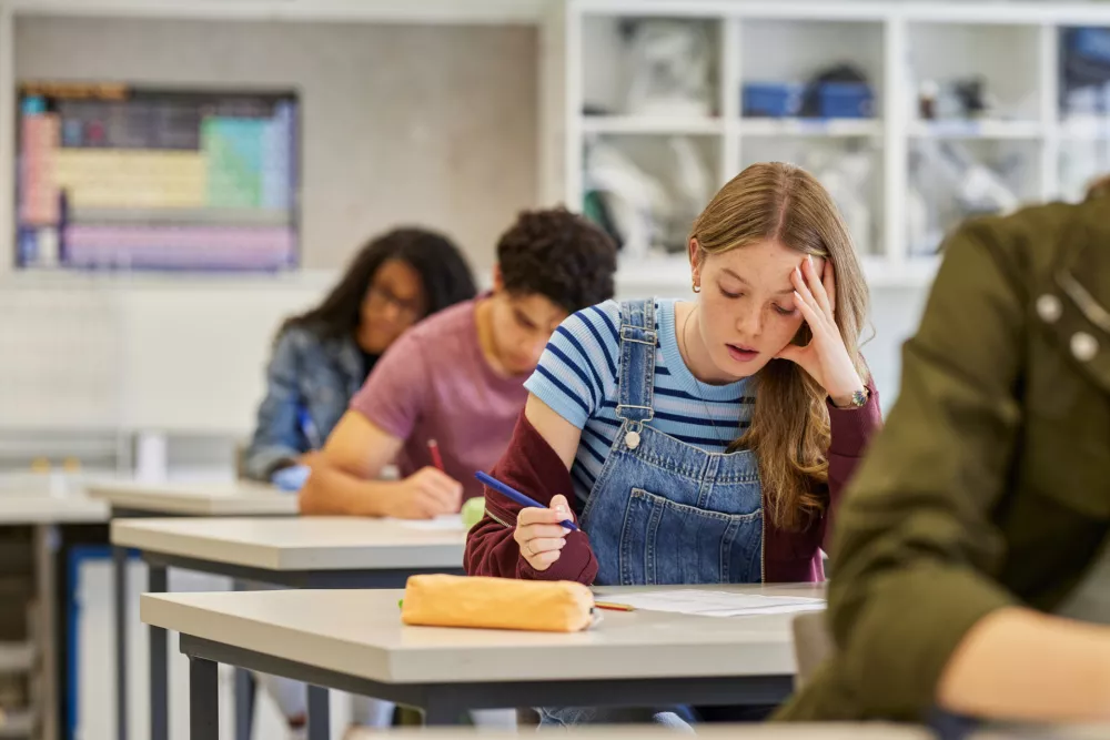 Stressed student holding head while taking exam in high school classroom. Tired girl showing signs of difficulty during test at secondary school with copy space. Classmates working silently while writing answers to the college entrance exam.