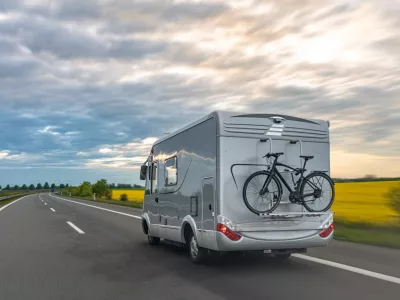 A gray motorhome drives on a highway with a bicycle mounted on the rear rack and open fields nearby. Useful for road trips, camping travel, route planning, motorhome tourism, and outdoor recreation content.