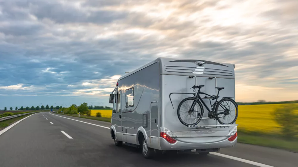 A gray motorhome drives on a highway with a bicycle mounted on the rear rack and open fields nearby. Useful for road trips, camping travel, route planning, motorhome tourism, and outdoor recreation content.