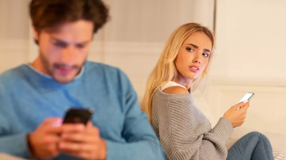 Infidelity. Wife Catching Cheating Husband Texting On Cellphone With Another Woman Sitting On Sofa At Home. Selective Focus