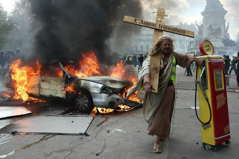 A protester dressed as Jesus Christ and holding a mock gas station dispenser walks past a burning car during a demonstration against the cost of living and rising fuel prices at Place de la Republique in Paris as part of nationwide protests called by the "Yellow Vests" movement, joined by French high school and university students, teachers and parents demanding better school conditions and government action, as well as labour unions, workers, farmers and retirees, France, October 17, 2026.  REUTERS/Abdul Saboor
