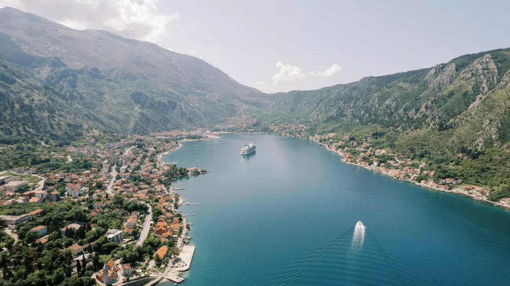 A panoramic aerial photograph of Kotor Bay in Montenegro, showing a cruise ship anchored in the calm blue waters, surrounded by lush green mountains and the historic town of Kotor along the shoreline.