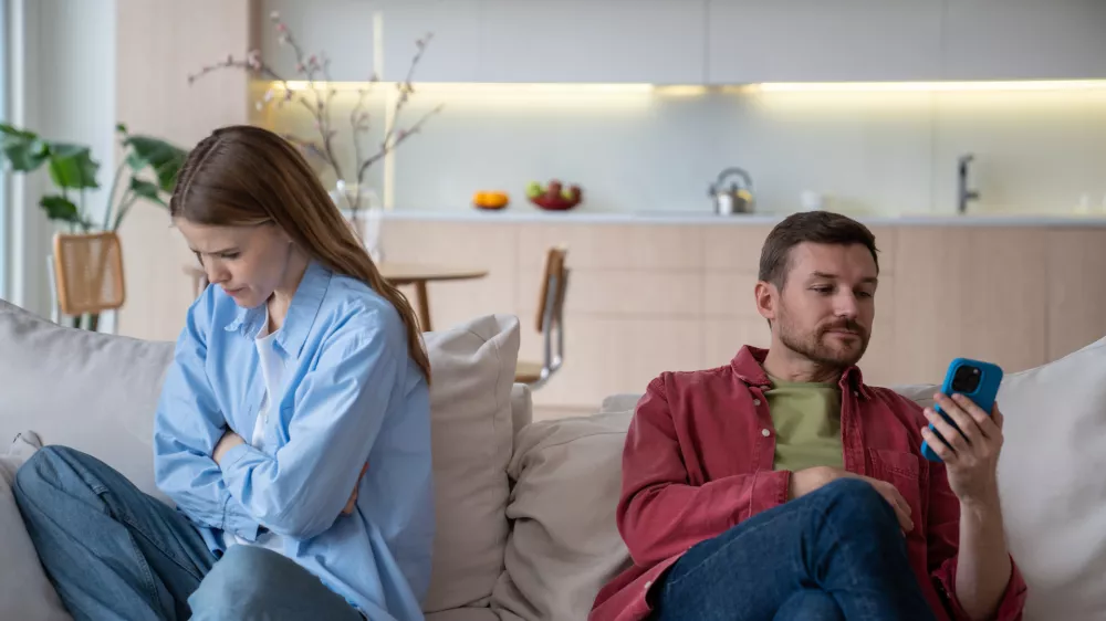 Jealous wife sad about husband messages in phone sitting on couch at home. Spouses ignoring each other. Upset woman turn out from man who looking at smartphone. Betrayal infidelity mistrust concept.