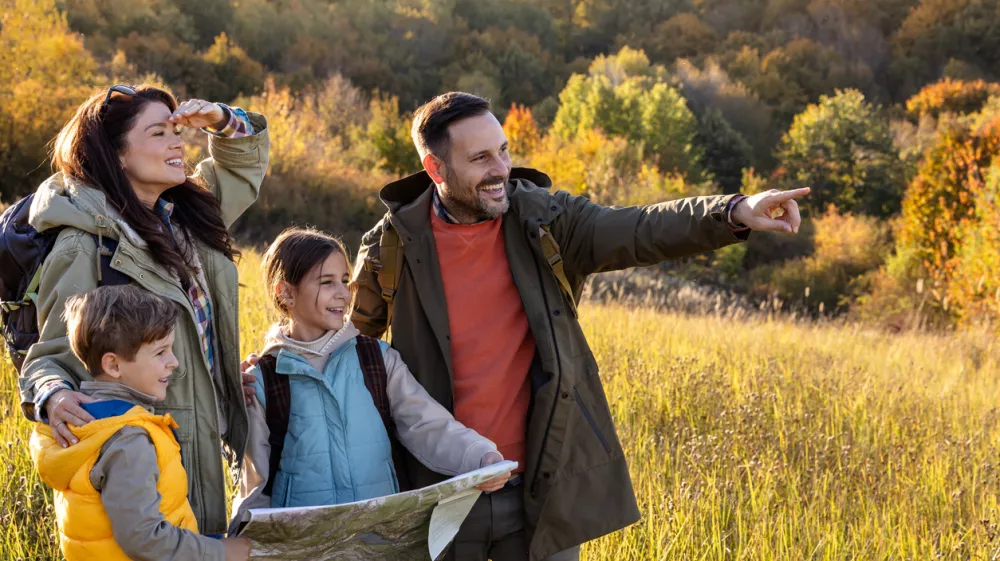 Family standing in sunny field while reading a map, children hold it and father points at viewpoint