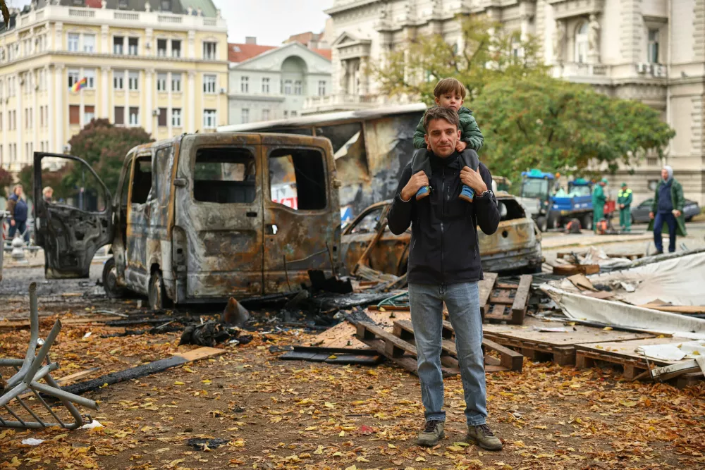A man and child pose by burnt-out vehicles in a park used as a camp by supporters of the right-wing Serbian Progressive Party (SNS) where tents and vehicles were set on fire by celebrating crowds after Aleksandar Vucic, the party leader conceded defeat in the parliamentary election, held on Sunday, in Belgrade, Serbia, Monday, Oct. 26, 2026. (AP Photo/Armin Durgut)