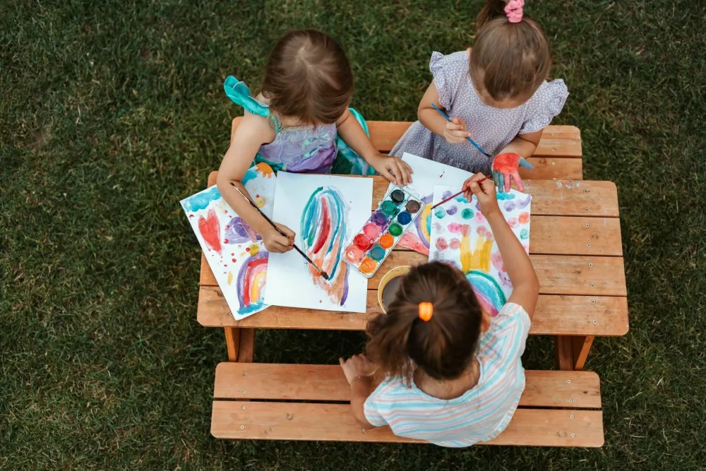 Three little girls enjoying a sunny day painting with watercolors at a wooden table in a garden / Foto: Vij