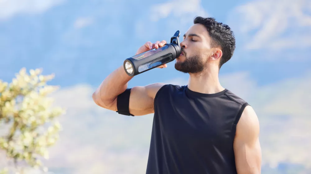 Fitness, blue sky and man drinking water from bottle to refresh and relax after workout, running or sports training. Cardio runner, exercise and thirsty young person with liquid drink for hydration