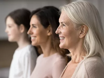 Happy attractive old 65s woman standing in row with young adult 30s daughter and little 7s granddaughter, smiling, look straight, side profile faces view. Three generations family portraits, close up